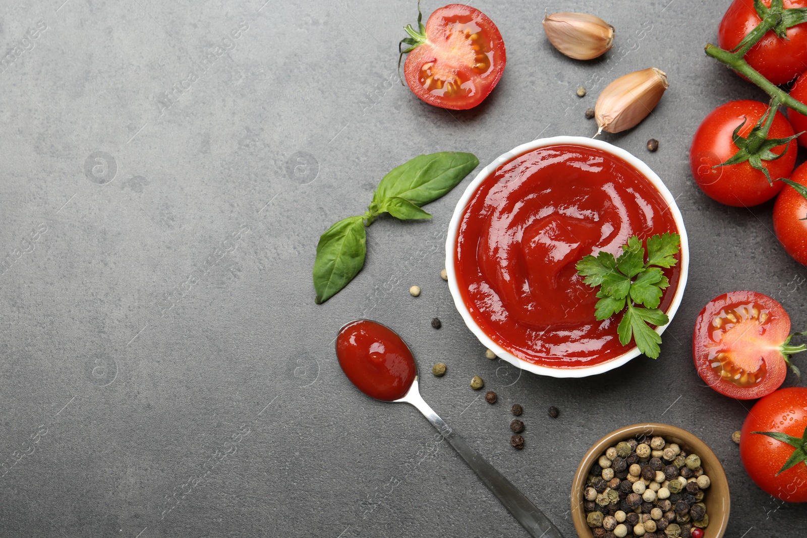 Photo of Delicious tomato ketchup in bowl, spices and products on grey textured table, flat lay. Space for text