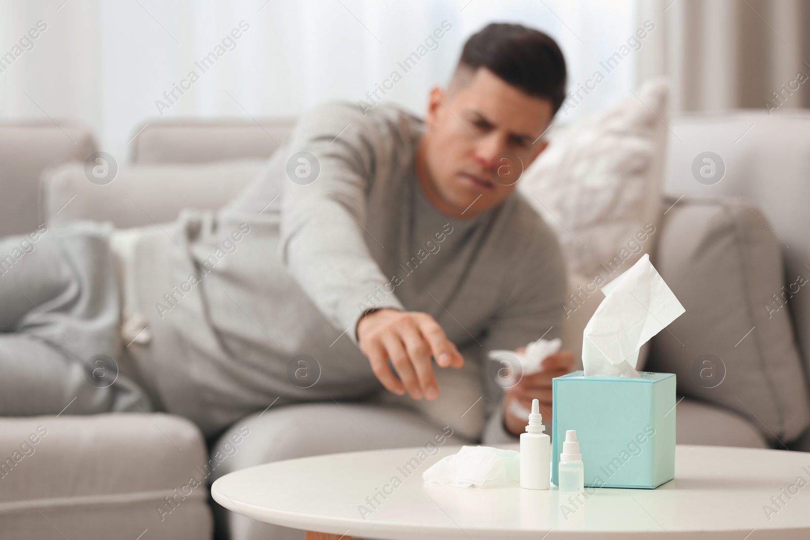 Photo of Ill man at home, focus on nasal spray, drops and box of tissues