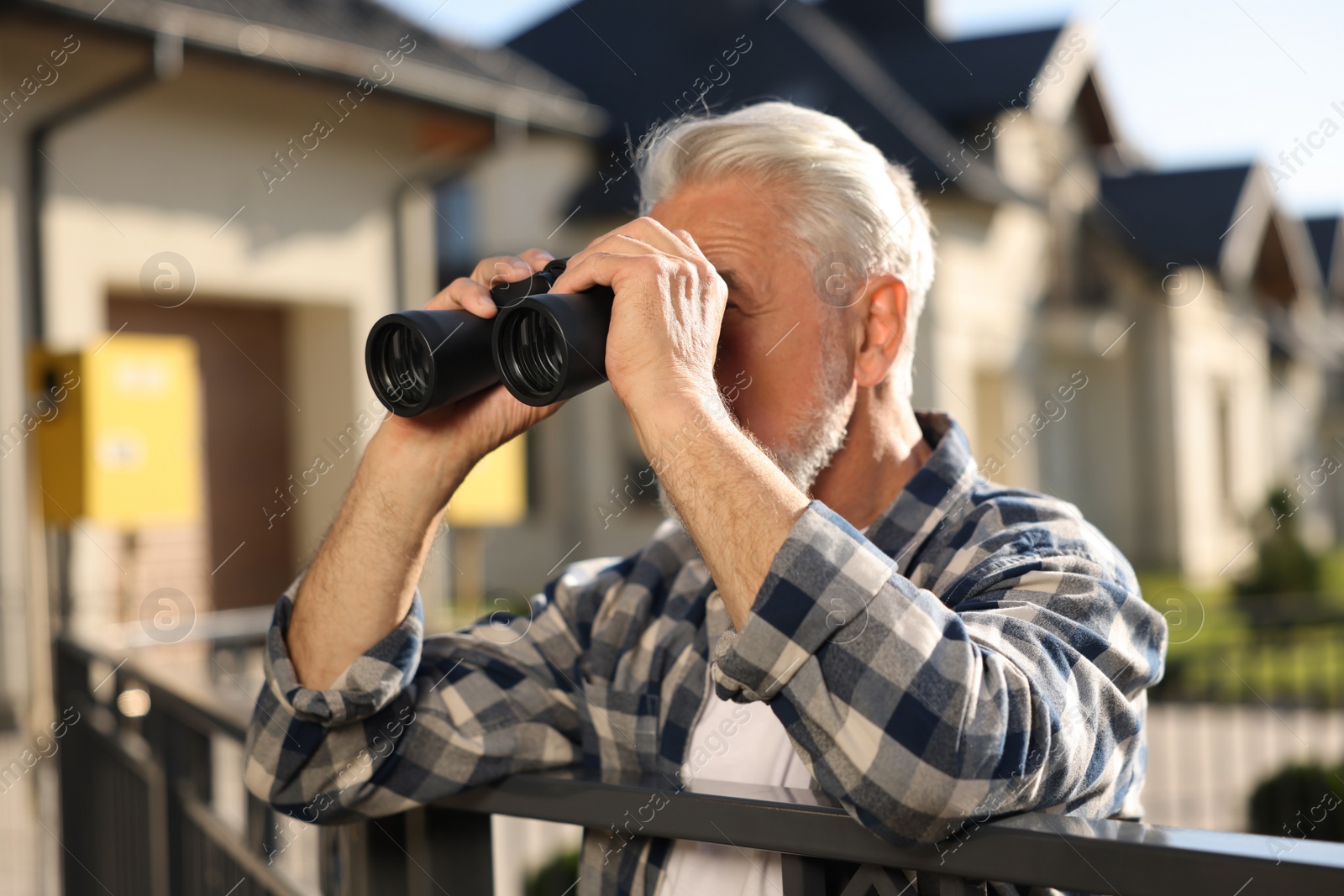 Photo of Concept of private life. Curious senior man with binoculars spying on neighbours over fence outdoors