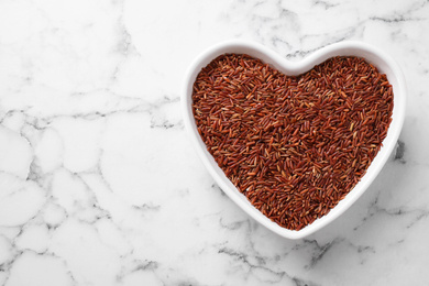 Photo of Brown rice in heart shaped bowl on white marble table, top view. Space for text