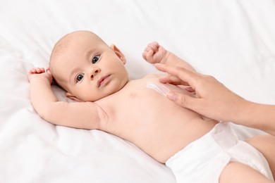 Photo of Woman applying body cream onto baby`s skin on bed, closeup