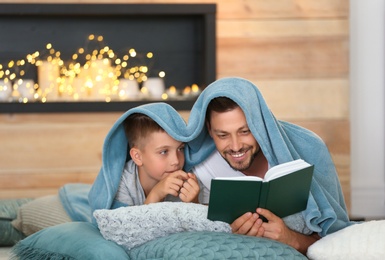 Photo of Dad and son reading interesting book near fireplace at home