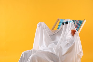 Photo of Person in ghost costume and sunglasses with glass of drink relaxing on deckchair against yellow background, space for text