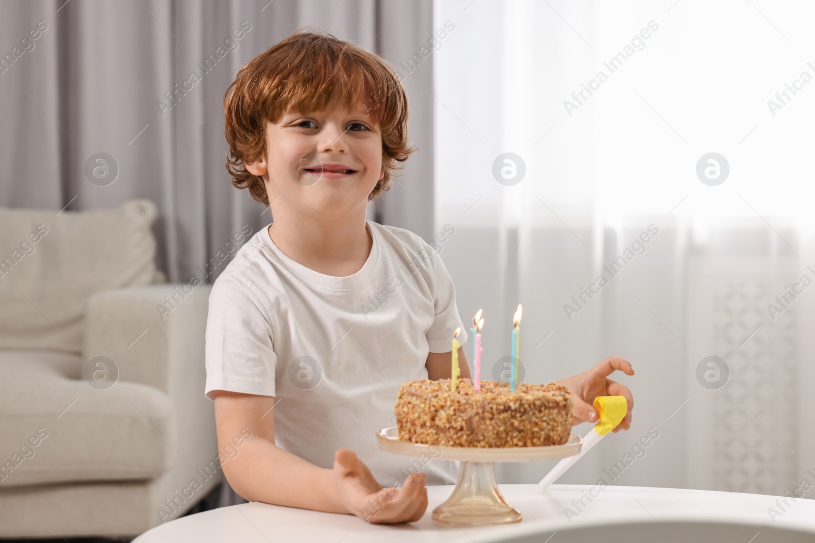 Photo of Birthday celebration. Cute boy with party blower at table with tasty cake indoors