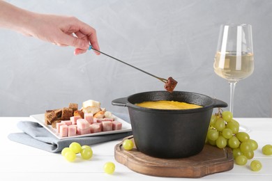 Photo of Woman dipping piece of bread into fondue pot with tasty melted cheese at white wooden table against gray background, closeup