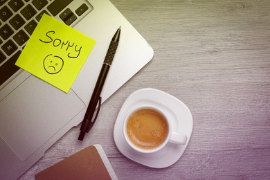 Image of Sticky note with word Sorry and drawn sad face attached to laptop on table. Workplace with cup coffee, flat lay