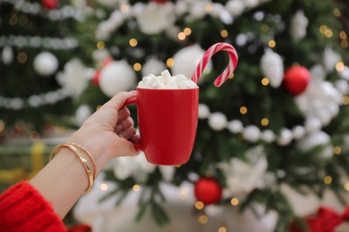 Photo of Woman with cup of delicious hot drink near Christmas tree indoors, closeup