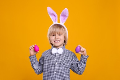 Photo of Happy boy in bunny ears headband holding painted Easter eggs on orange background