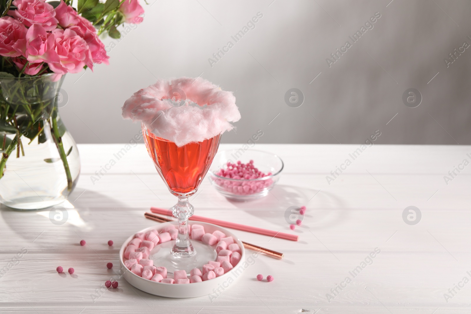 Photo of Cotton candy cocktail in glass, marshmallows, vase with pink roses and straws on white wooden table against gray background, space for text