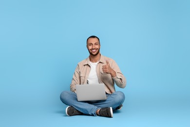 Smiling young man with laptop showing thumbs up on light blue background