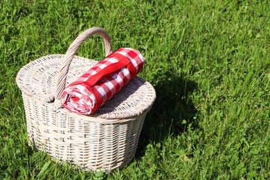 Photo of Rolled checkered tablecloth with picnic basket on green grass outdoors, space for text