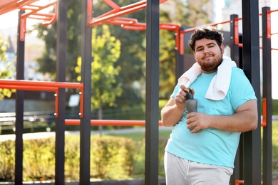 Young overweight man with bottle and towel on sports ground