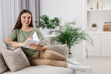 Photo of Beautiful young woman reading book on sofa in room with green houseplants