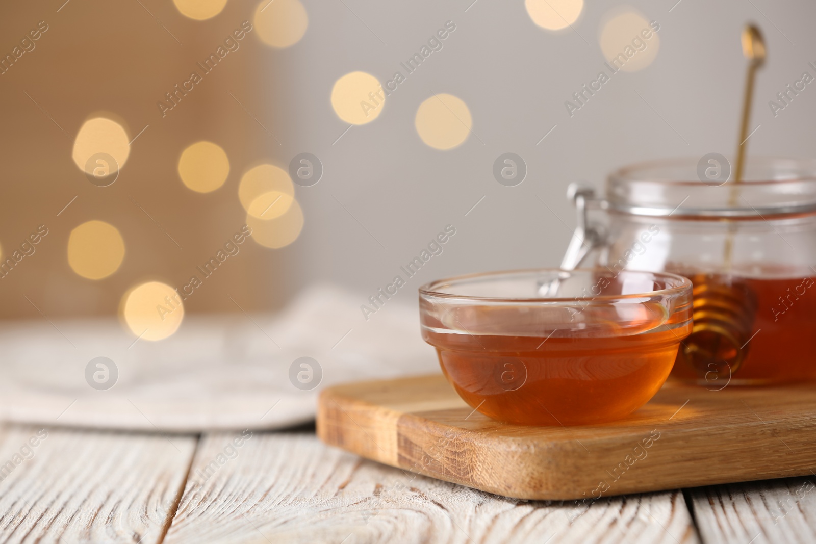 Photo of Delicious honey in glass dishware on white wooden table against blurred lights, closeup. Space for text