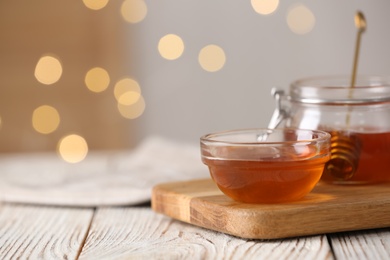 Photo of Delicious honey in glass dishware on white wooden table against blurred lights, closeup. Space for text