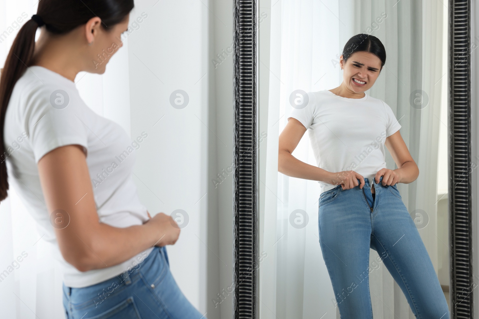 Photo of Young woman trying to put on tight jeans near mirror indoors