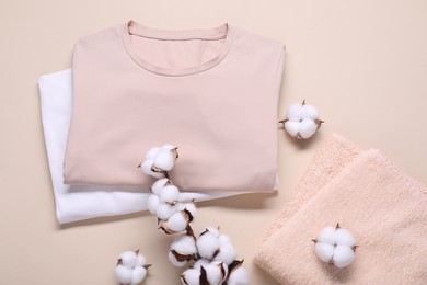 Photo of Cotton branch with fluffy flowers, t-shirts and terry towel on beige background, flat lay