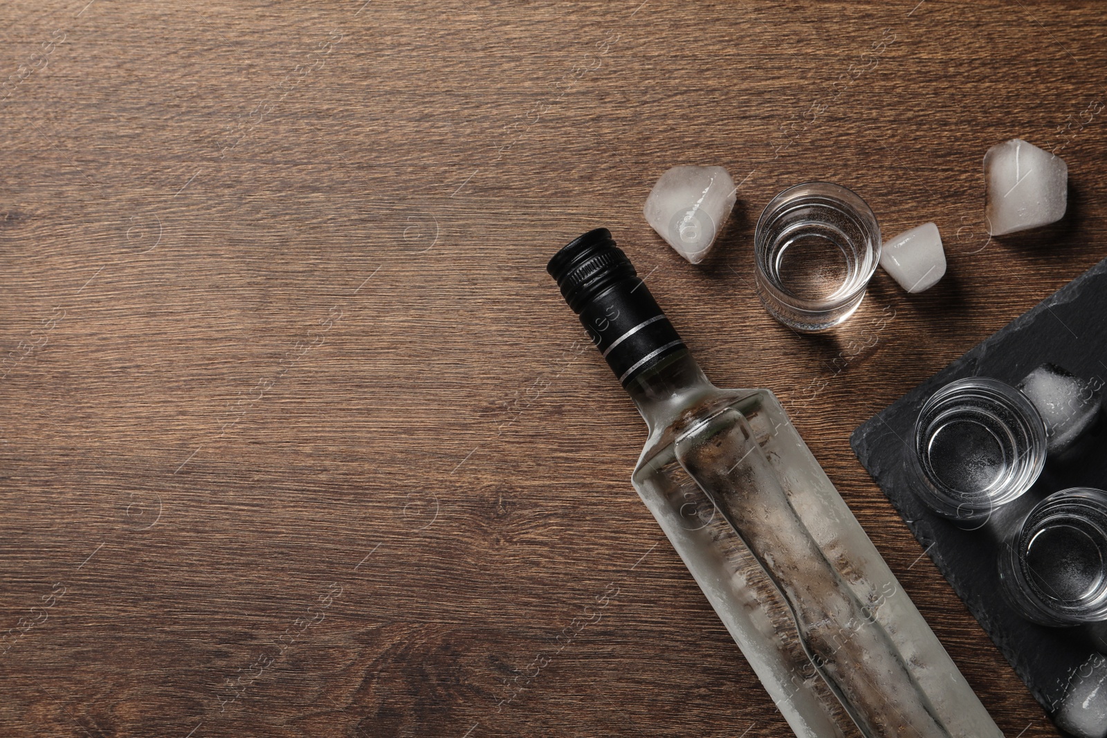 Photo of Bottle of vodka, shot glasses and ice cubes on wooden table, flat lay. Space for text