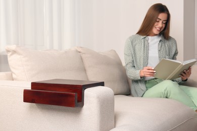Photo of Wooden armrest table on sofa in room. Woman reading book at home, selective focus