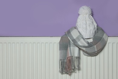 Knitted hat and scarf on heating radiator near violet wall