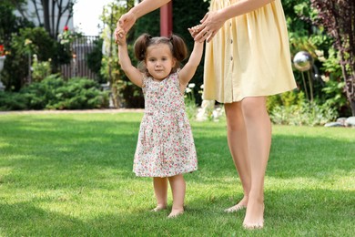 Photo of Mother supporting baby girl while she learning to walk on green grass in park