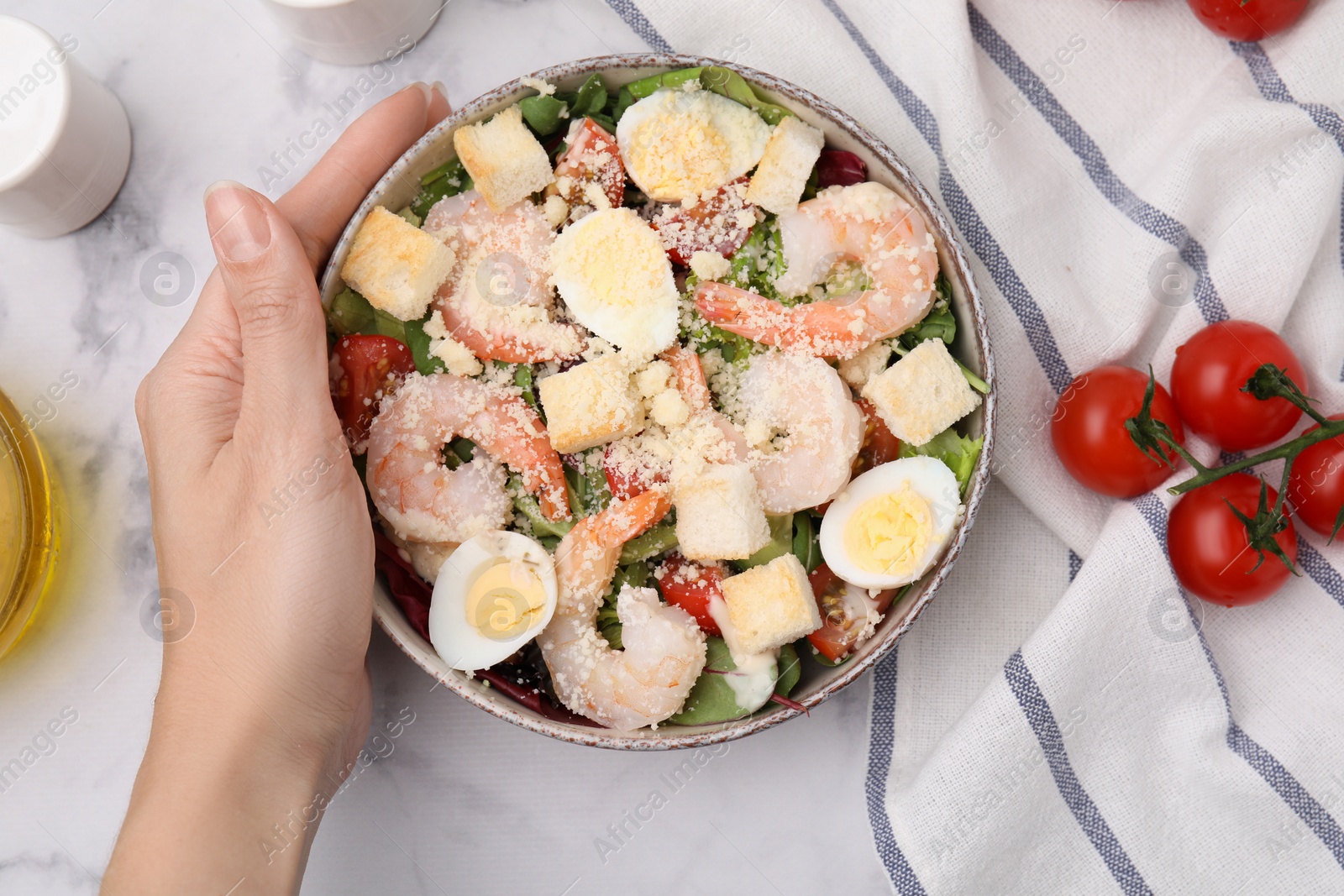 Photo of Woman holding bowl of delicious Caesar salad with shrimps at white marble table, top view