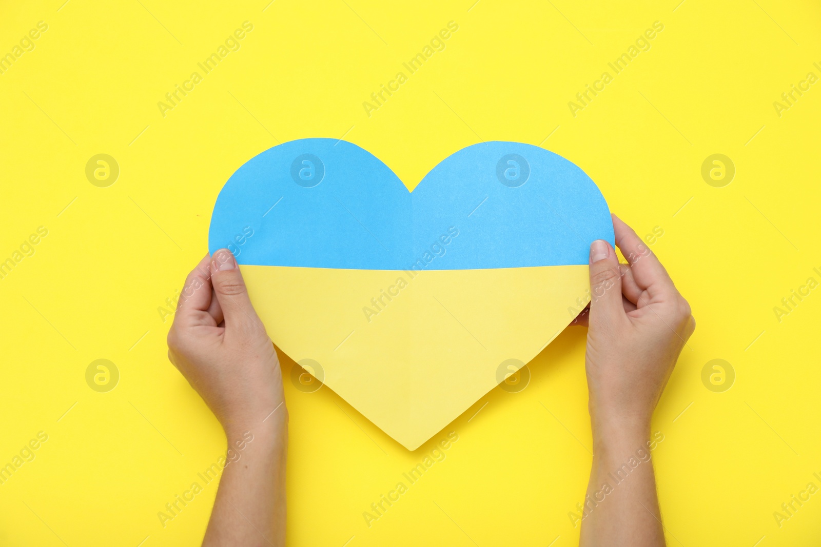 Photo of Woman holding paper heart in colors of Ukrainian flag on yellow background, top view