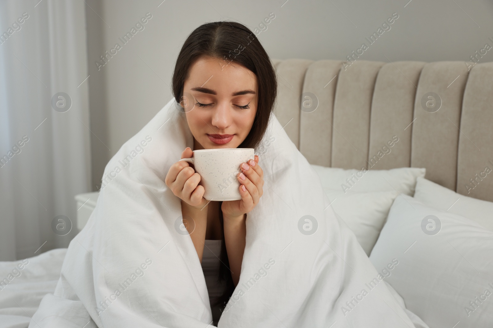 Photo of Beautiful young woman with cup of drink wrapped in blanket at home