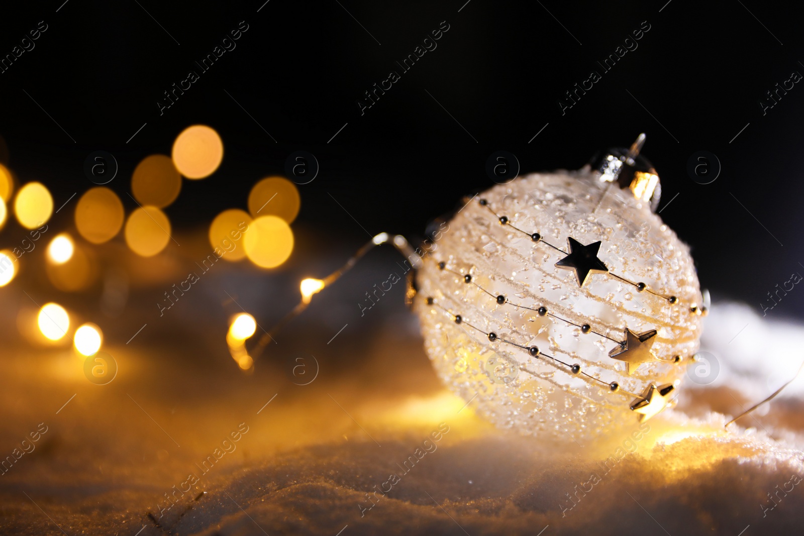 Photo of Christmas ball and fairy lights on white snow outdoors. Space for text