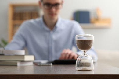 Photo of Hourglass with flowing sand on desk. Man using calculator indoors, selective focus