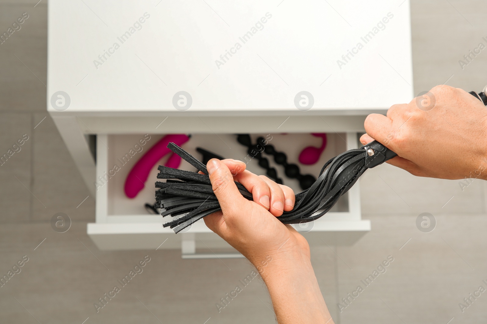Photo of Man holding black leather whip over drawer with other sex toys in room, above view