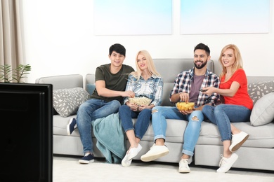Young people with snacks watching TV on sofa at home