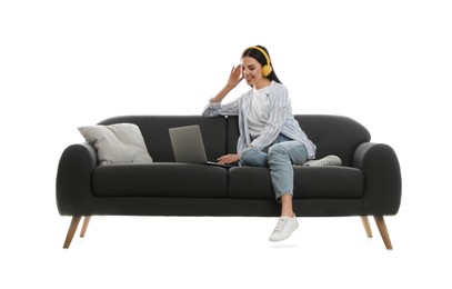 Young woman with laptop listening to music on comfortable grey sofa against white background