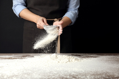 Photo of Woman sifting wheat flour at table against black background, closeup