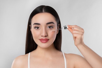 Young woman applying essential oil onto face on light grey background, closeup