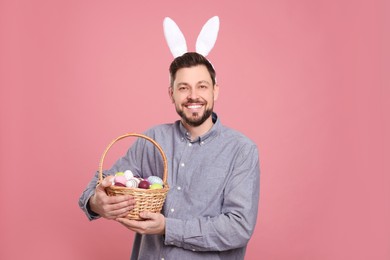Photo of Happy man in bunny ears headband holding wicker basket with painted Easter eggs on pink background
