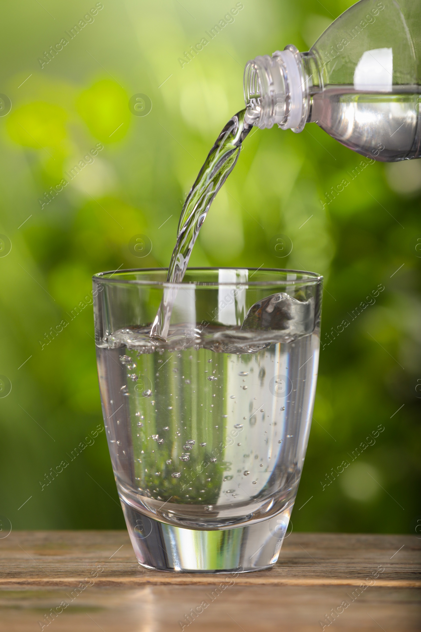 Photo of Pouring water from bottle into glass on wooden table outdoors, closeup