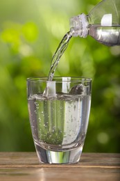 Photo of Pouring water from bottle into glass on wooden table outdoors, closeup