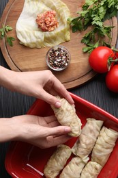 Woman putting uncooked stuffed cabbage roll into baking dish at black table, top view