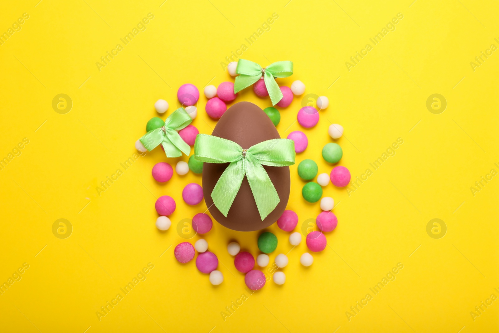 Photo of Delicious chocolate egg, light green bows and colorful candies on yellow background, flat lay