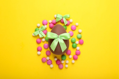 Photo of Delicious chocolate egg, light green bows and colorful candies on yellow background, flat lay
