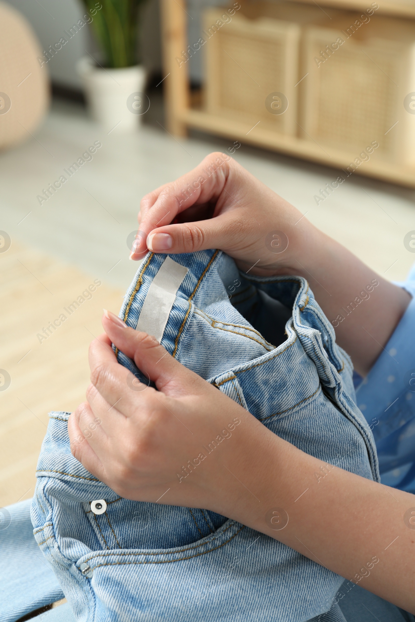 Photo of Woman holding jeans with blank clothing label at home, closeup