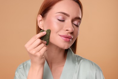 Young woman massaging her face with jade gua sha tool on pale orange background