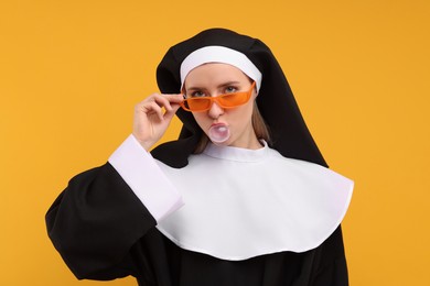 Woman in nun habit and sunglasses blowing bubble gum against orange background