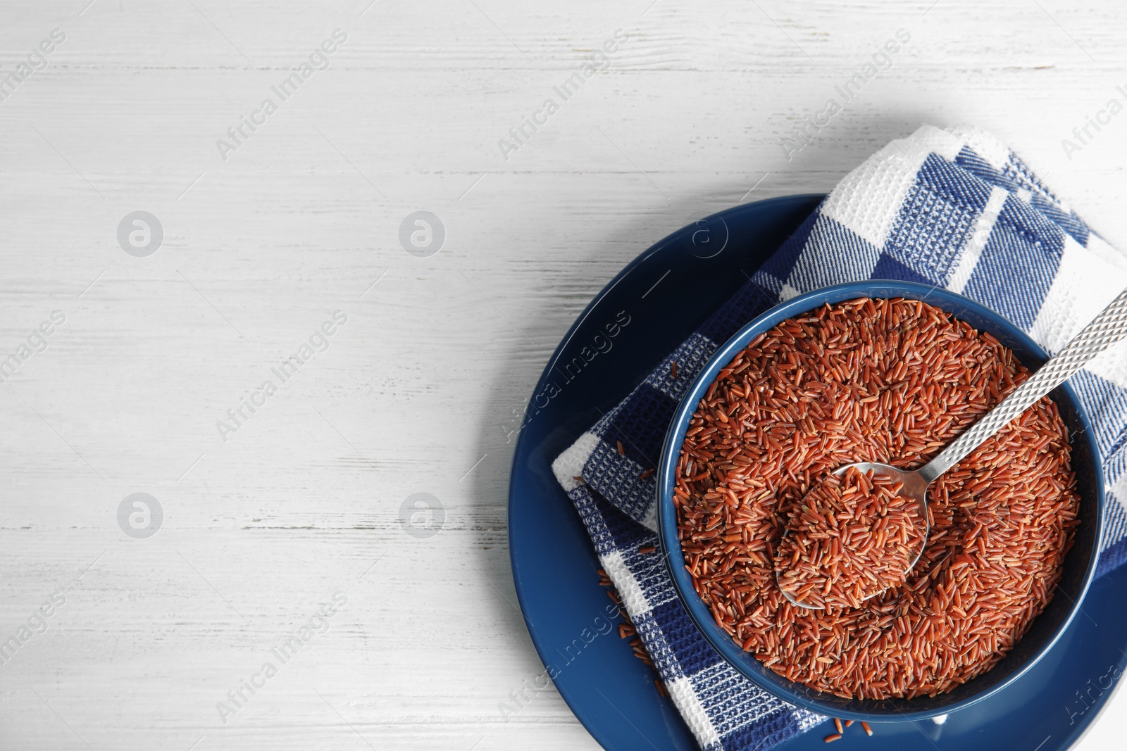 Photo of Bowl with uncooked red rice and spoon on white table, top view. Space for text