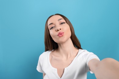 Beautiful young woman blowing kiss while taking selfie on light blue background