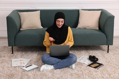 Photo of Muslim woman in hijab using video chat on laptop near sofa indoors