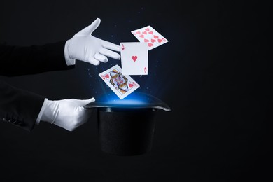 Magician showing trick with cards and top hat on black background, closeup