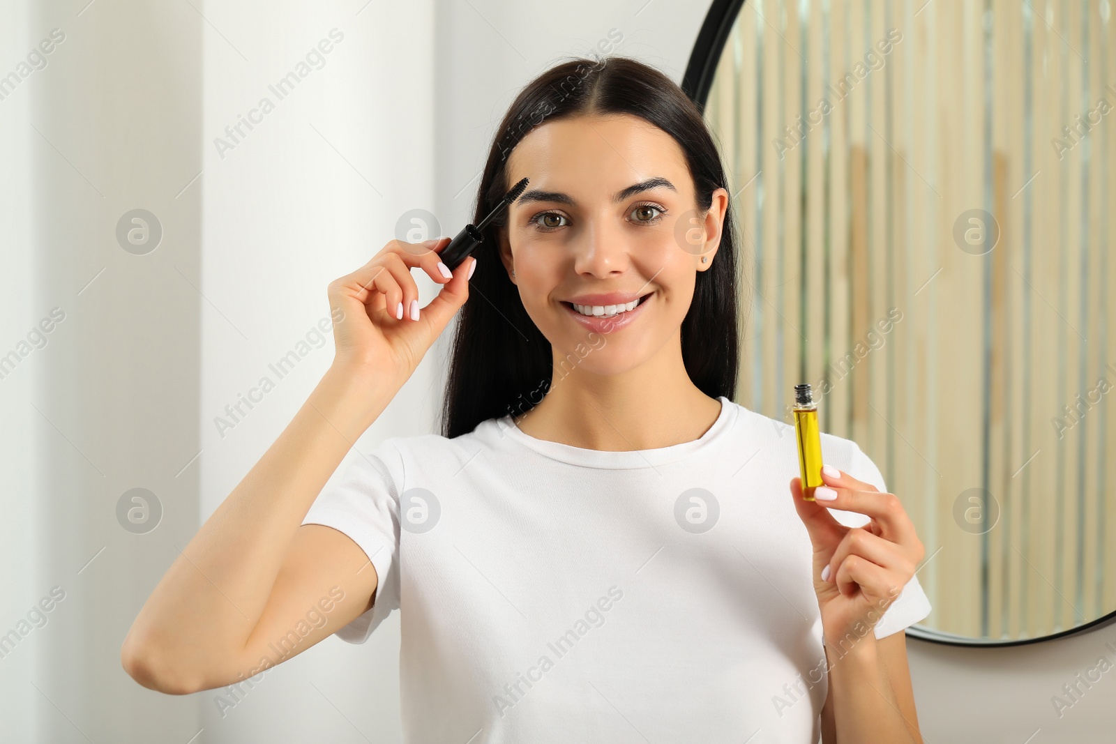 Photo of Young woman applying oil onto eyelashes near mirror indoors