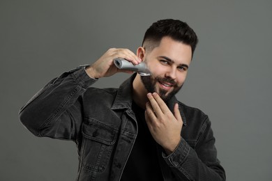 Handsome young man trimming beard on grey background